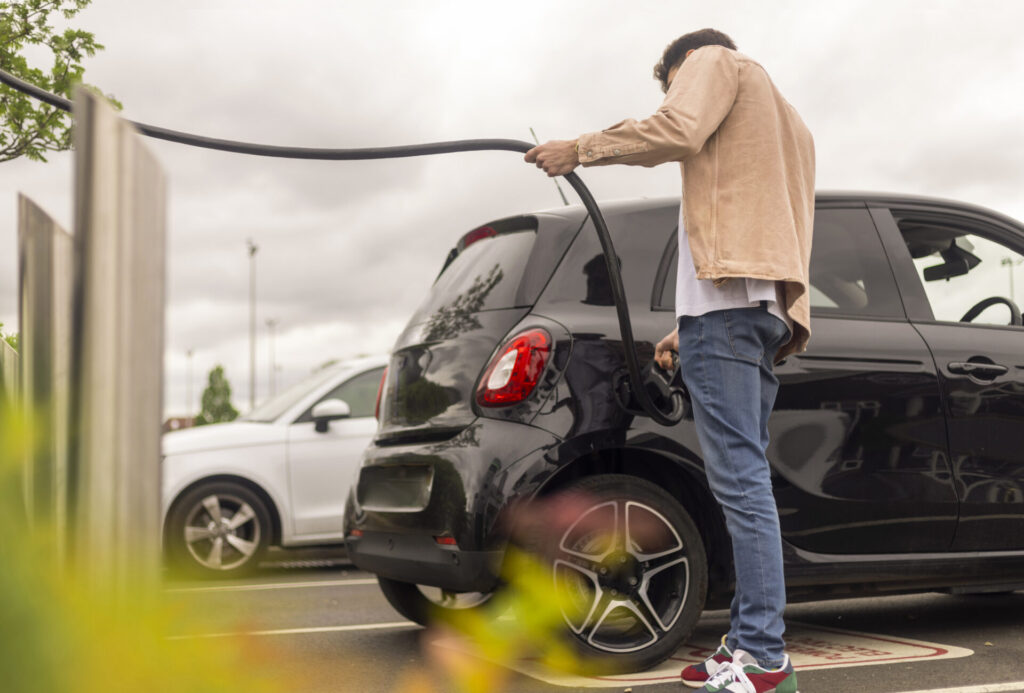 A rear view shot of young adult plugging a charger into his car in a car park. He is wearing casual clothing.