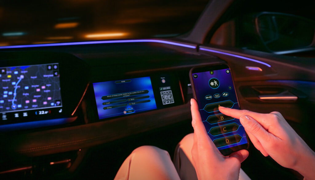 The AirConsole gaming platform app and the game Who Wants to Be a Millionaire? run on the front passenger display in an Audi. A person is holding a smartphone in front of the display. They use the smartphone as a controller to play the game and select answer options.